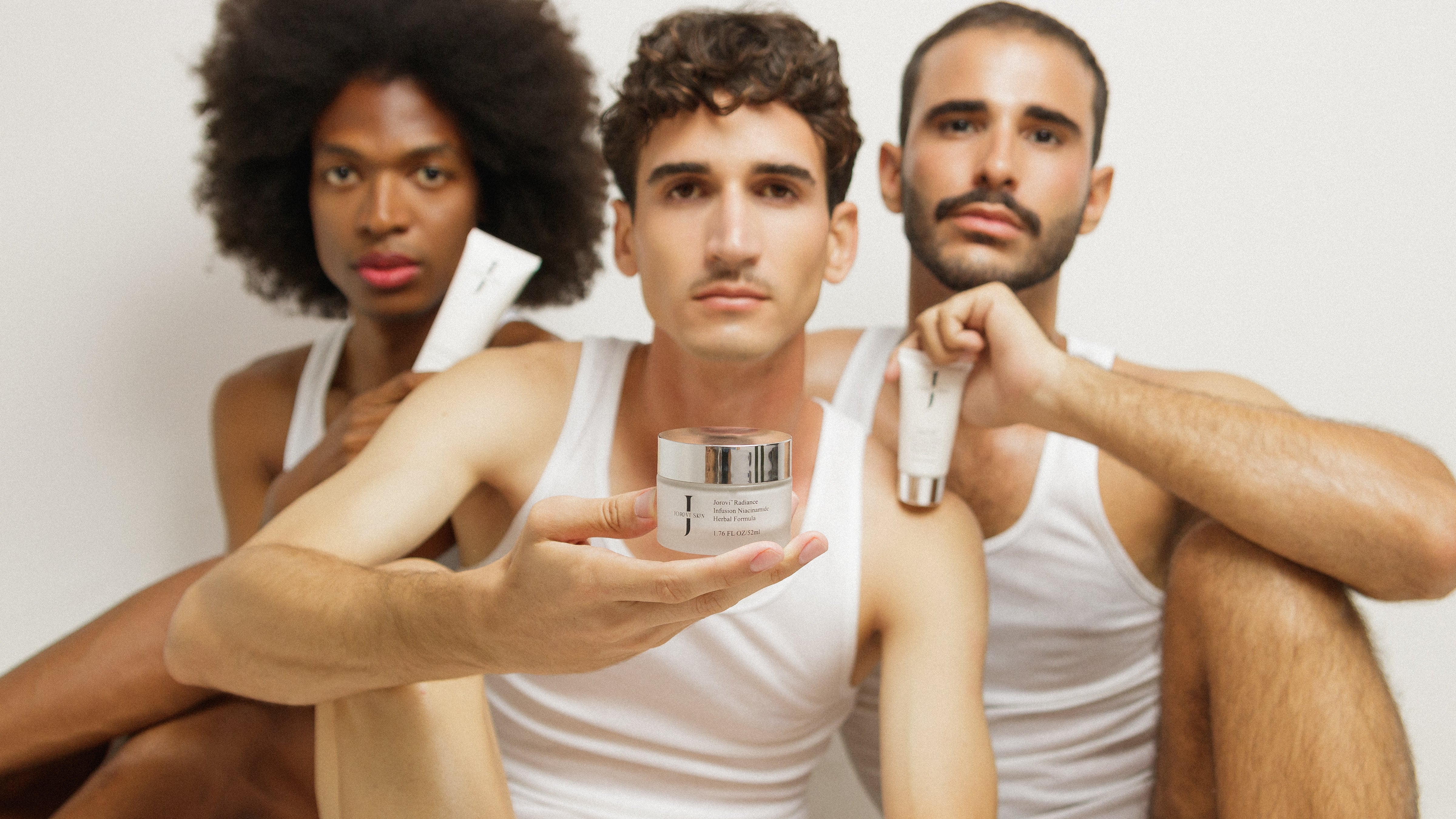 Three men of diverse backgrounds sitting together, holding Jorovi Skin products, including a moisturizer jar and skincare tubes, while wearing white tank tops, showcasing a skincare routine for men.