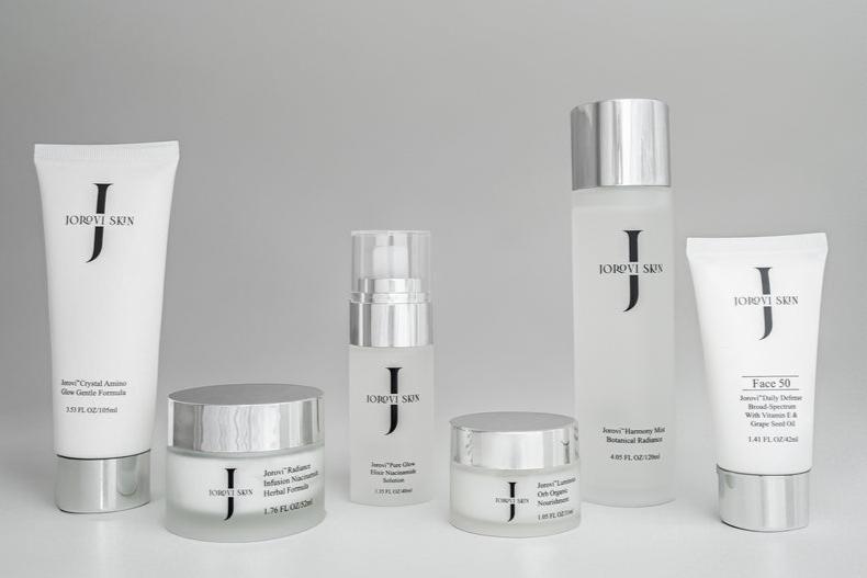 A woman with glowing, healthy skin applies Jorovi Moisturizer to her cheek while holding the product jar, demonstrating proper skincare technique.