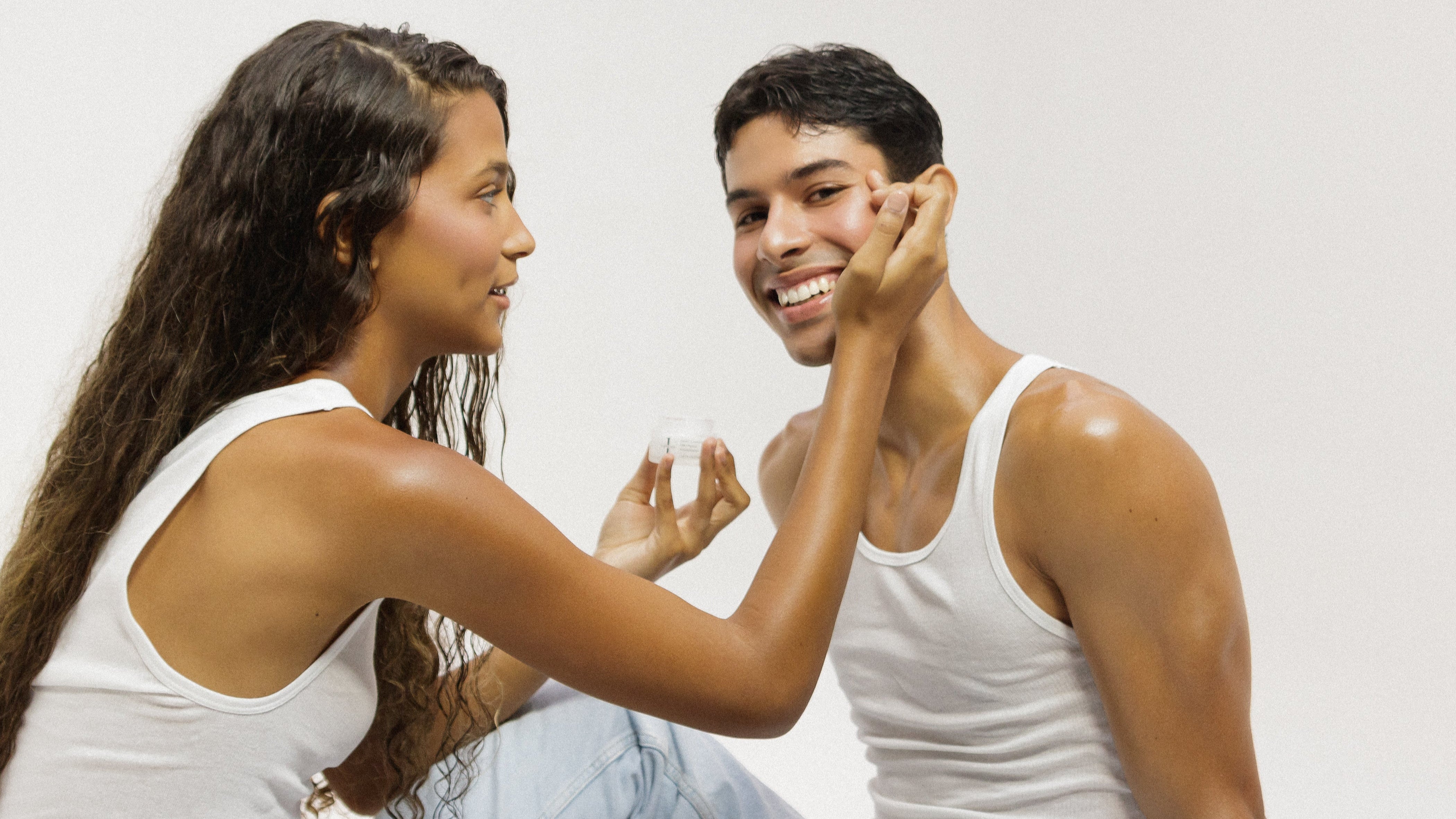 Smiling woman applying Jorovi Skin cream to a man's cheek, both wearing white tank tops and sitting together, showcasing skincare application and care.